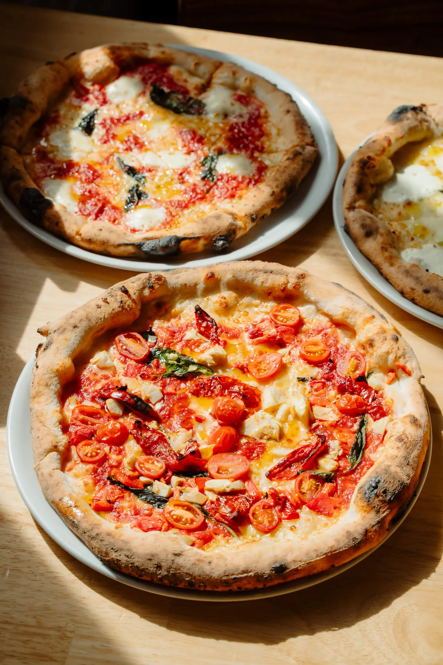Three pizzas with melted cheese, tomatoes, and garnish on a wooden table.